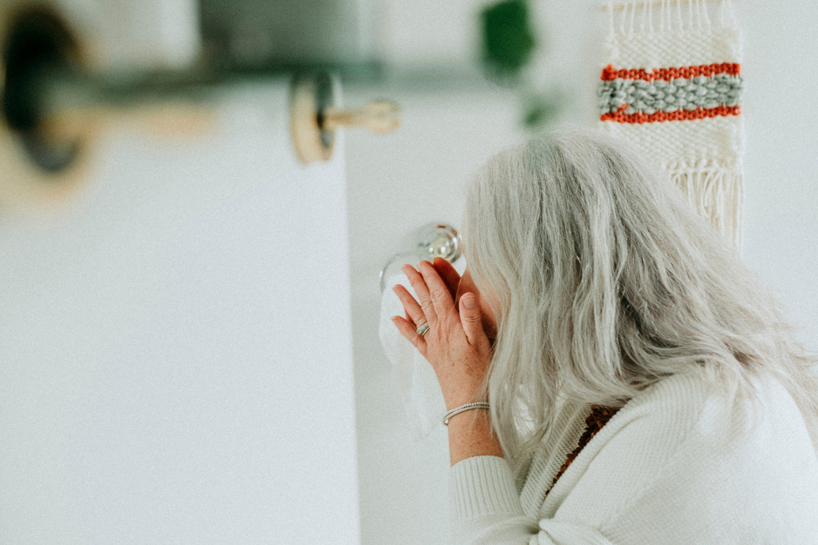 A woman with long gray hair, wearing a white knit sweater, is seen from behind drinking from a glass mug in a bright, softly lit bathroom. A bohemian-style woven wall hanging with red, orange, and gray stripes hangs on the wall next to green trailing ivy.